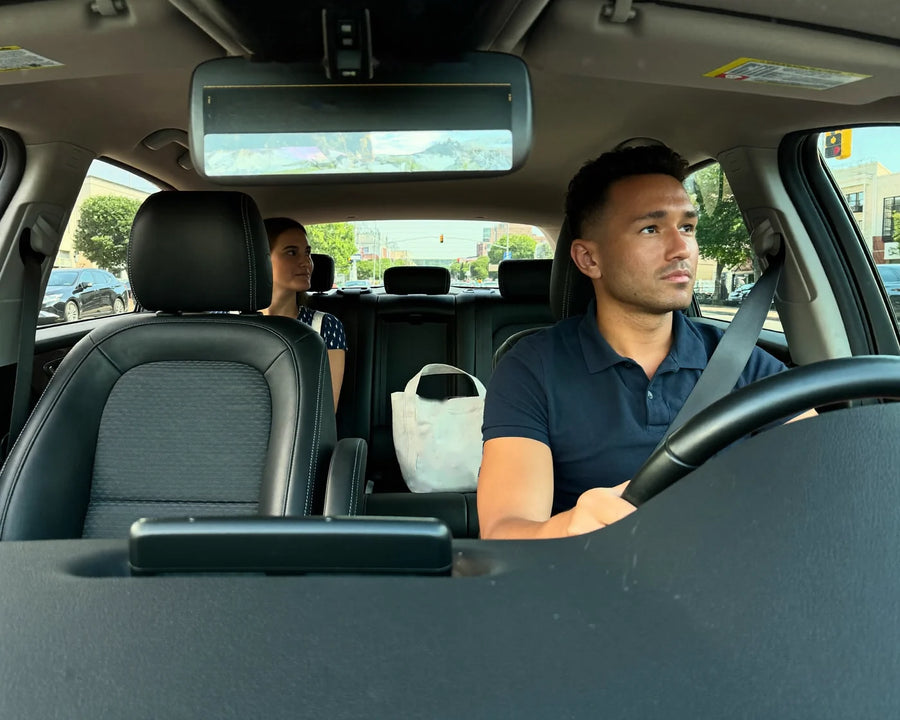 A rideshare driver at the wheel with a passenger seated behind him, city street and traffic lights visible through the windows.